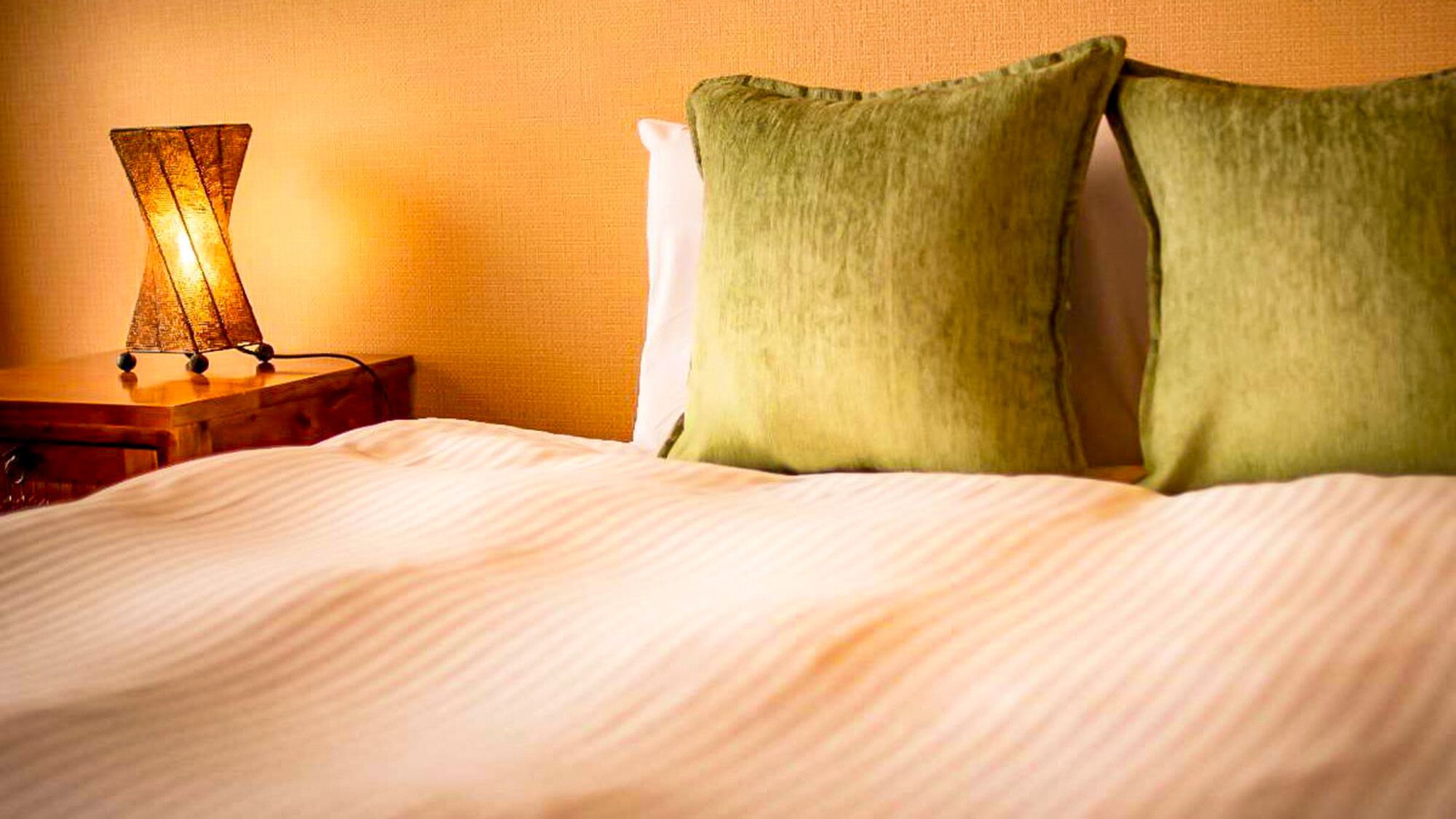 A neatly made bed with a white comforter and two green pillows, a bedside lamp, and a wooden nightstand.