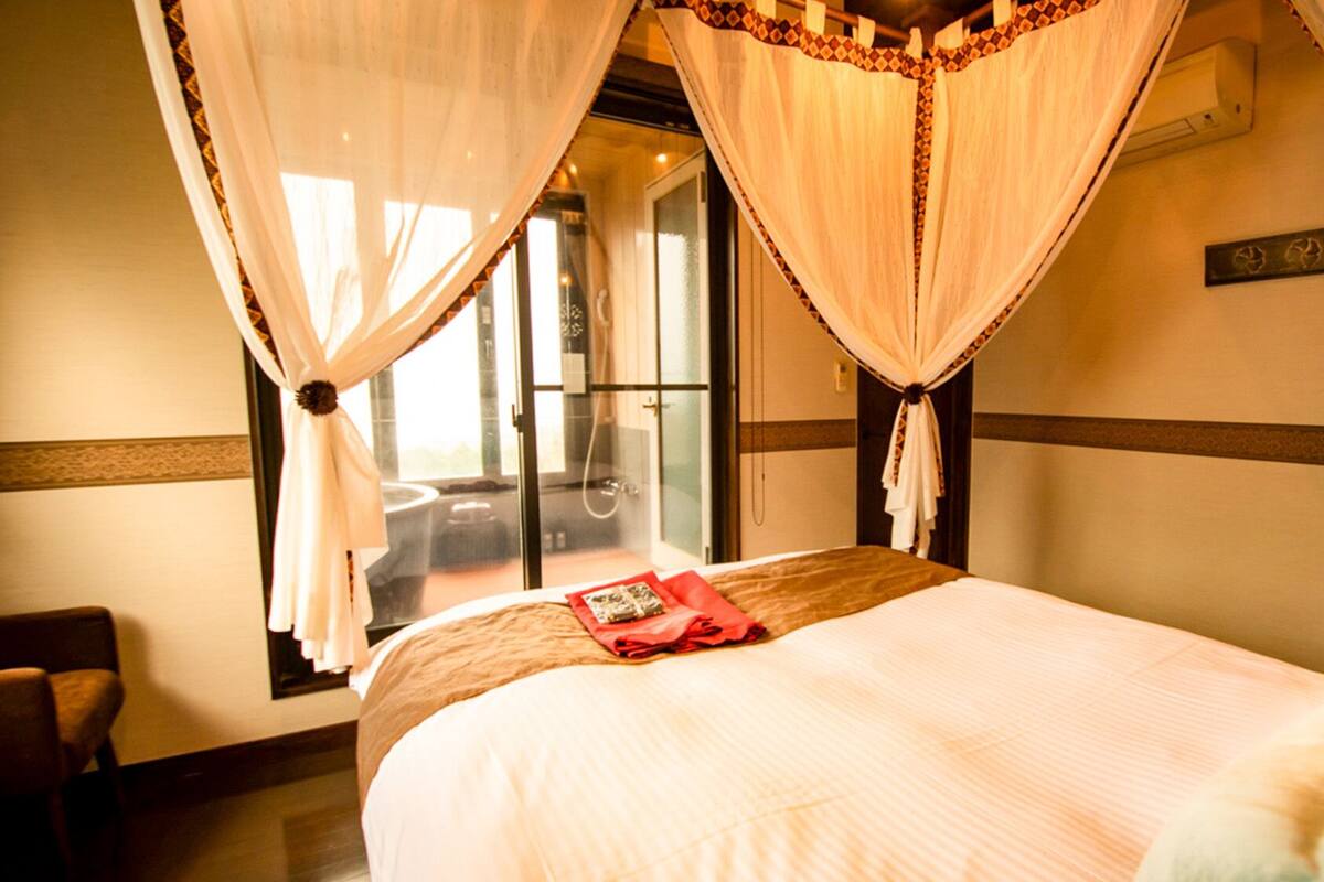 A hotel room with a bed, a chair, a window with curtains, and a sliding door leading to a balcony.