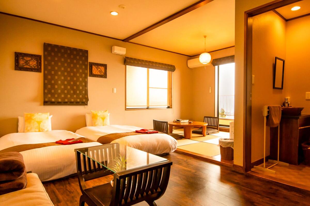 A hotel room with two beds, a dining table, a chair, a small table, and a mirror.