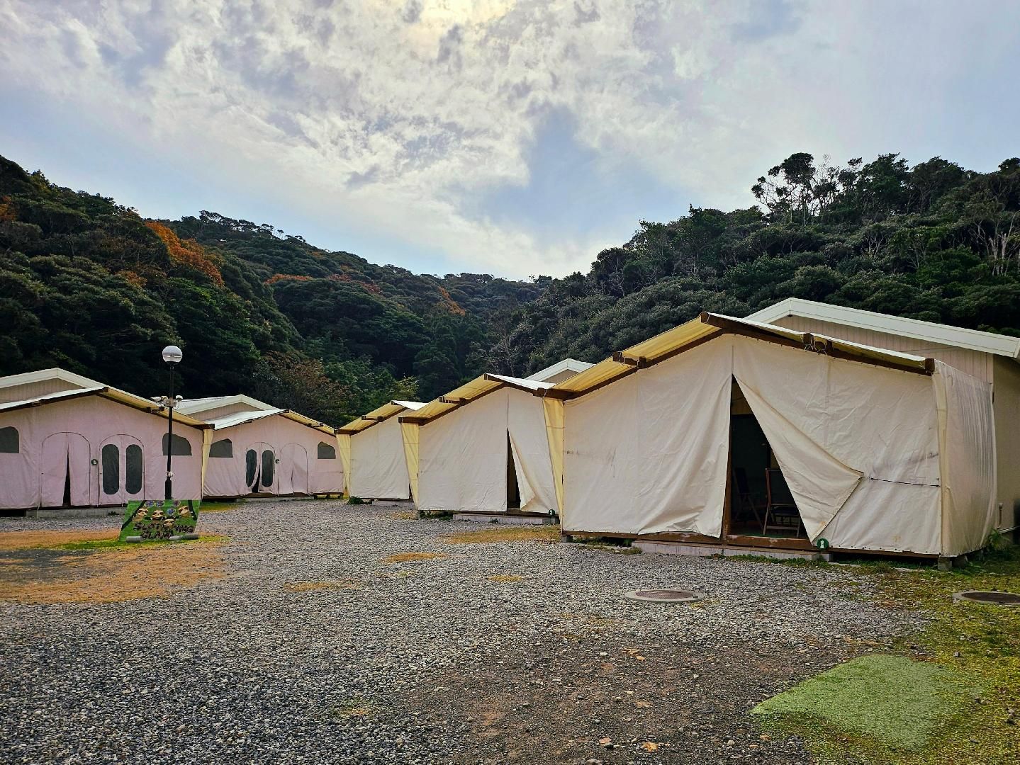 A row of simple, tent-like structures with a gravel ground, surrounded by dense forest.