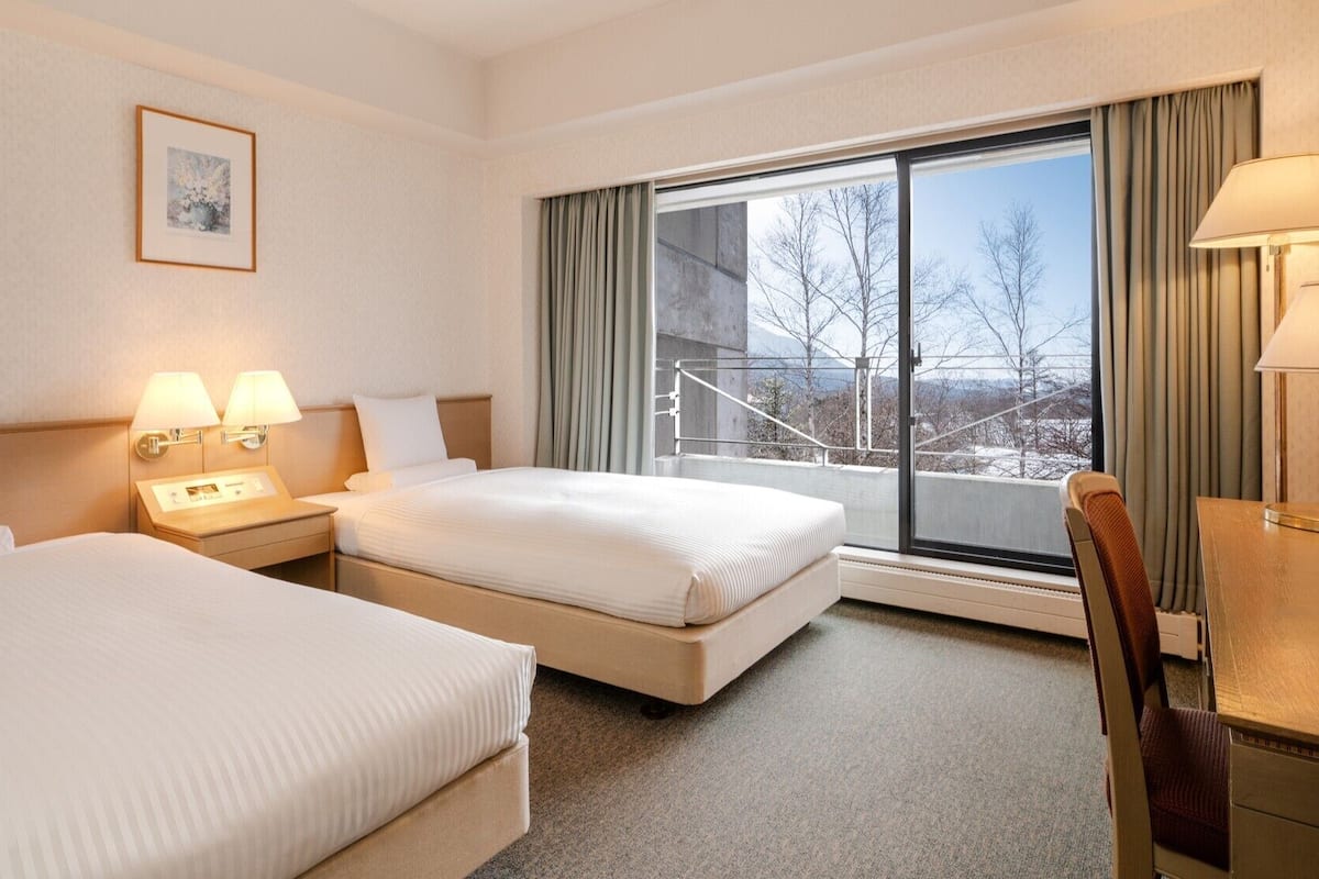 A hotel room with two beds, a desk, a chair, a window with a view, and a framed picture on the wall.