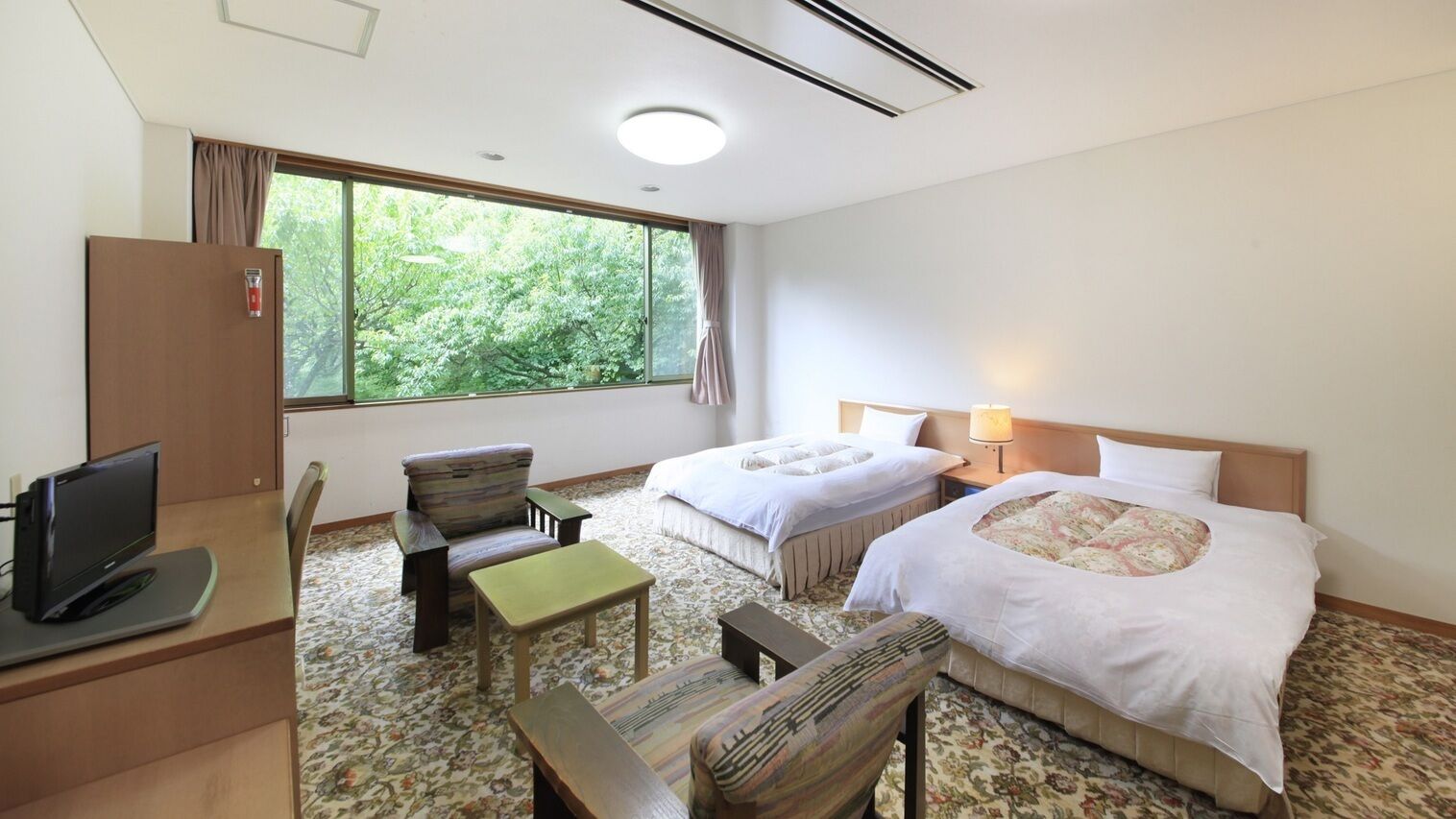 A hotel room with two beds, a desk with a computer, a chair, a small table, and a window with a view of trees.