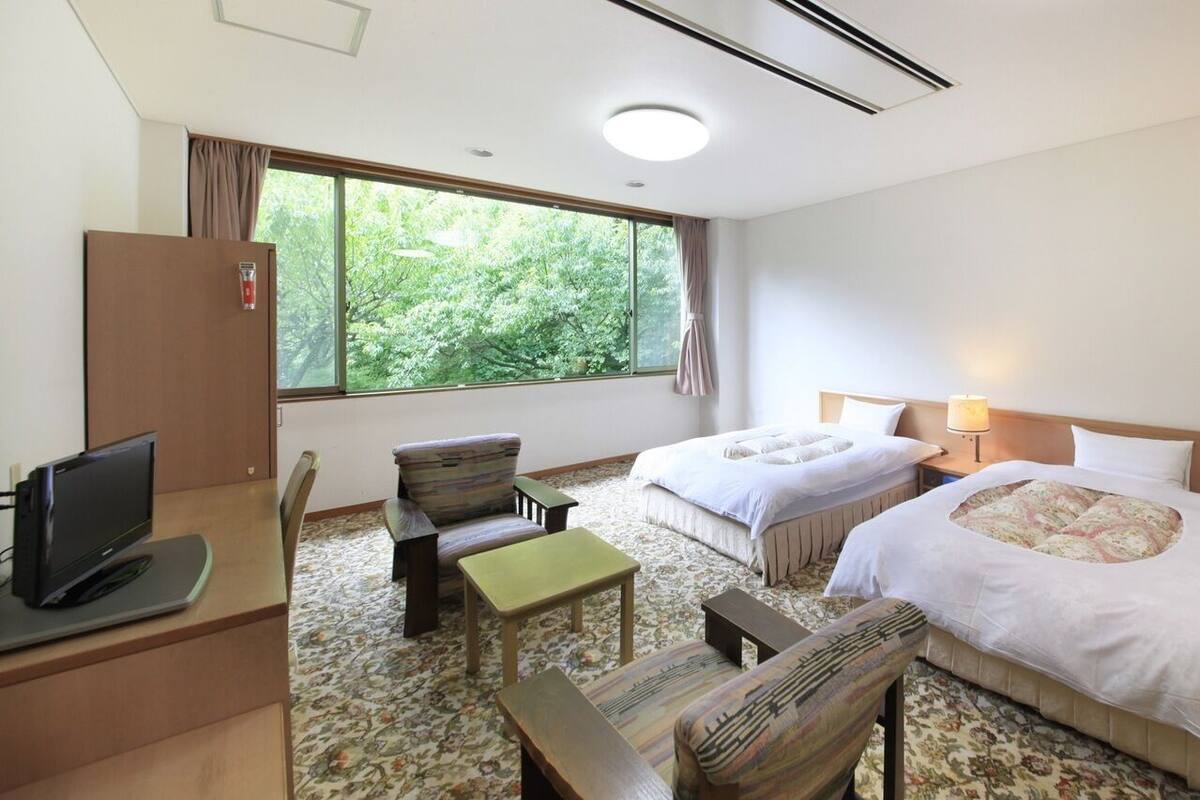 Habitación de hotel con dos camas, un escritorio con computadora, una silla, una mesita y ventana con vista a árboles.