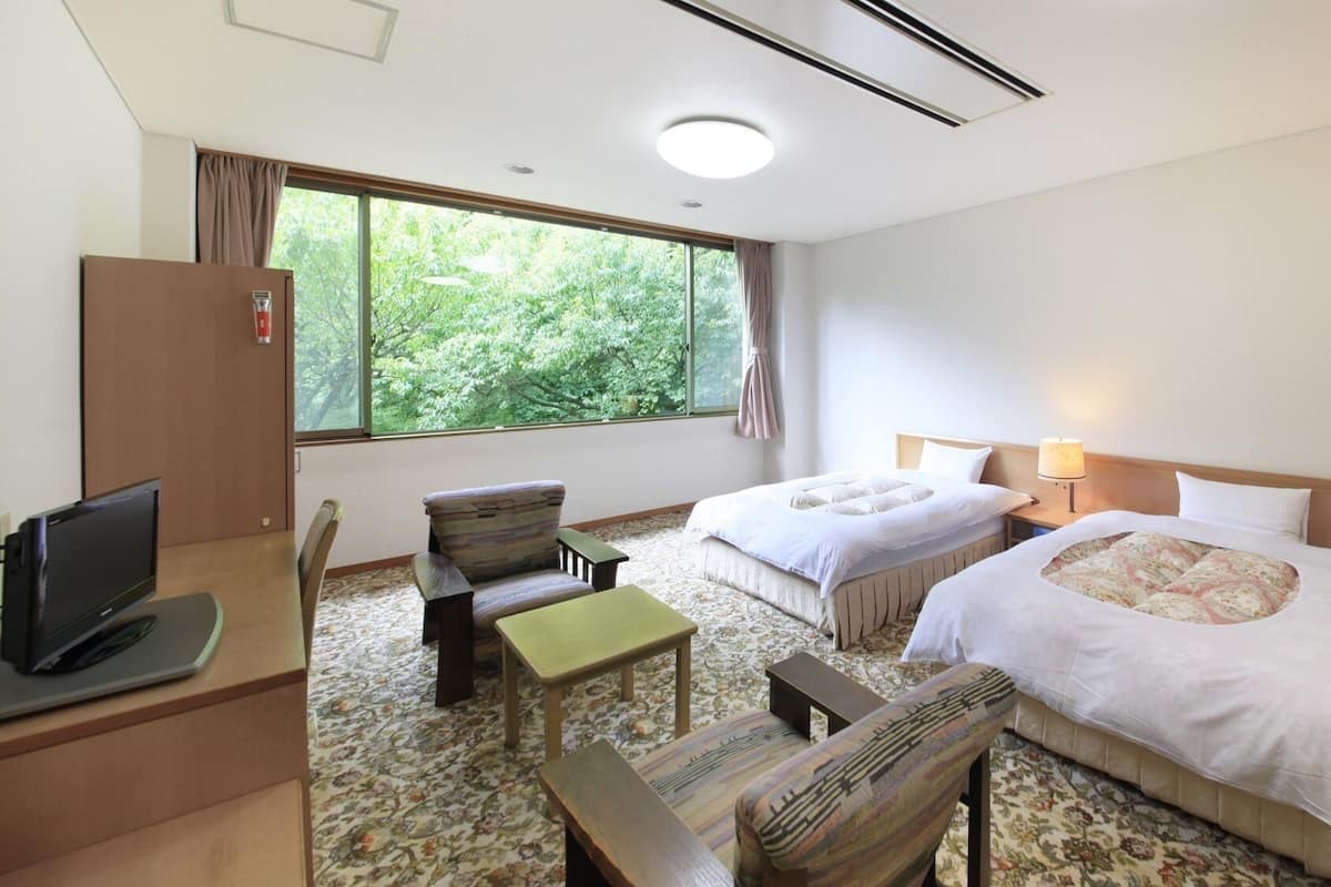 Habitación de hotel con dos camas, un escritorio con computadora, una silla, una mesita y una ventana con vista a árboles.