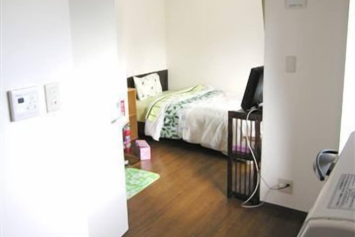 Ein Zimmer mit einem Bett, einem Schreibtisch und einem Stuhl. Es gibt ein Fenster mit Vorhängen, eine Tür und eine Wand-Klimaanlage.