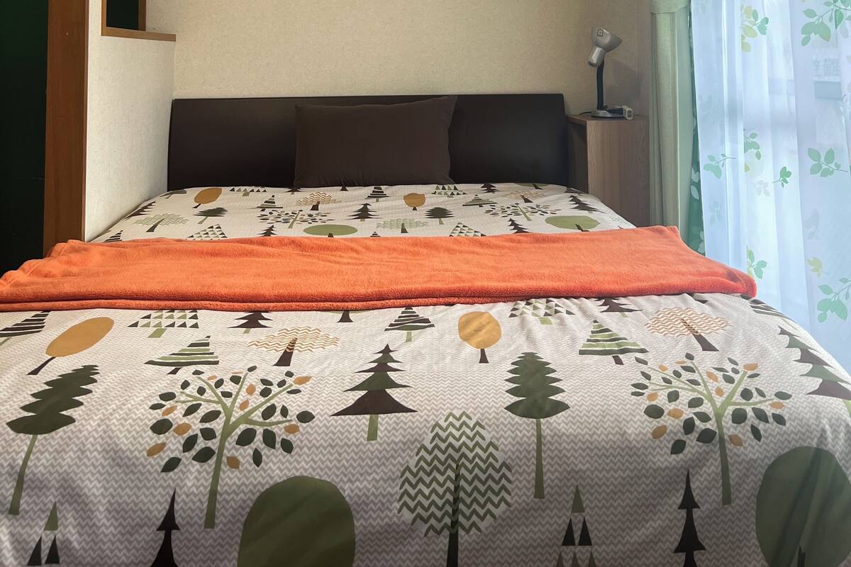Ein Bett mit gemustertem Überwurf, ein Nachttisch mit Lampe und ein Fenster mit blumigen Vorhängen.
