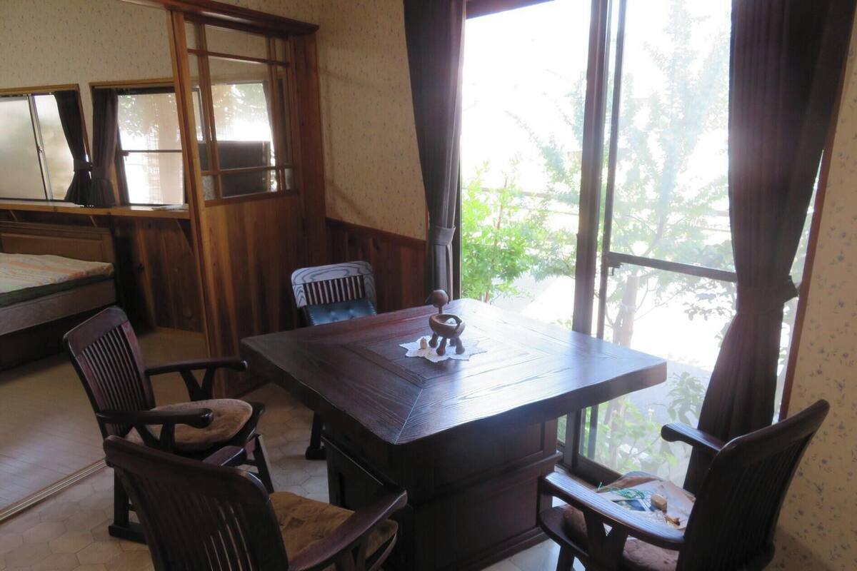 A room with a wooden table, two chairs, a bed, and large windows with curtains.