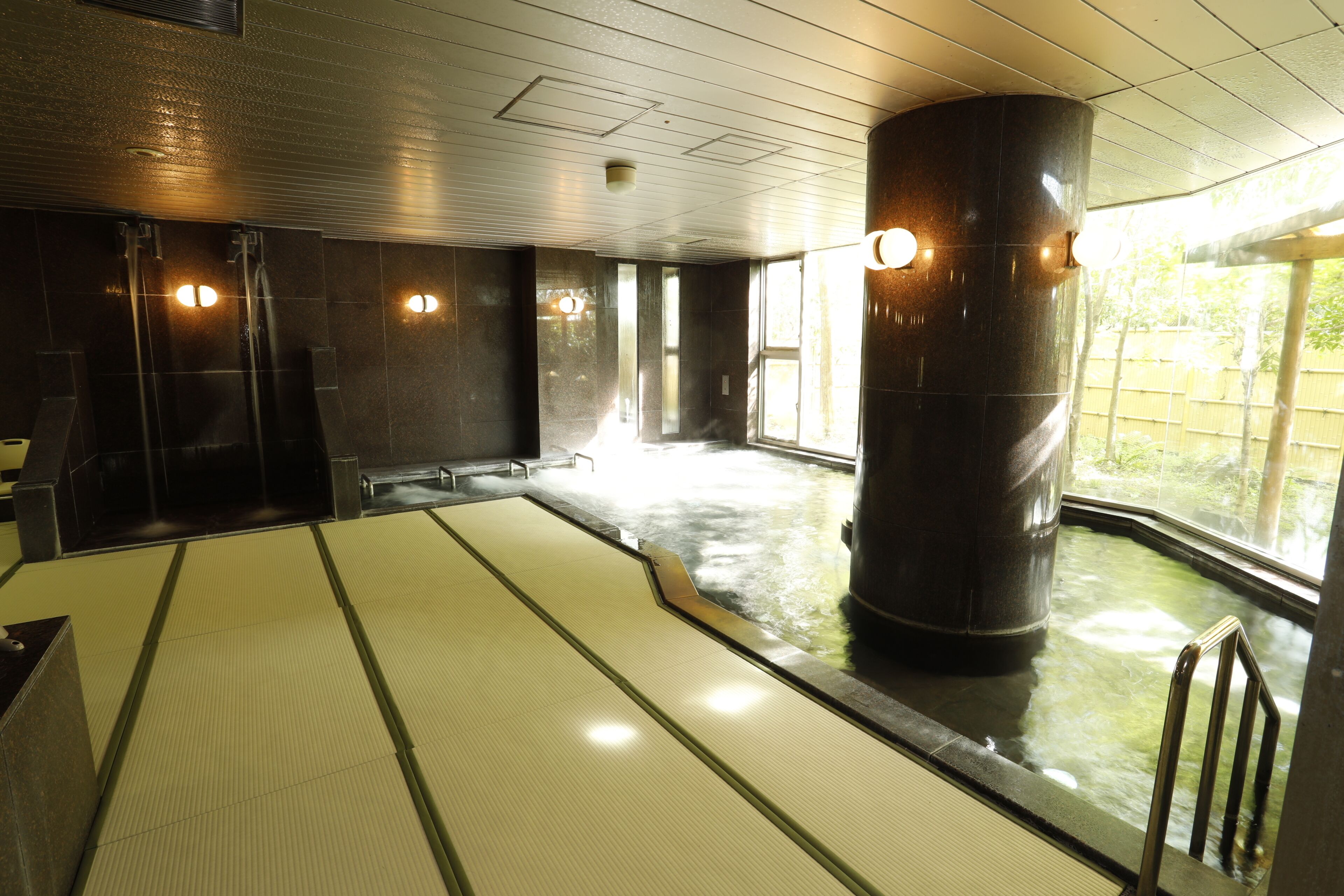 A large indoor pool with a tiled floor, a central column, and a view of the outdoors through large windows.