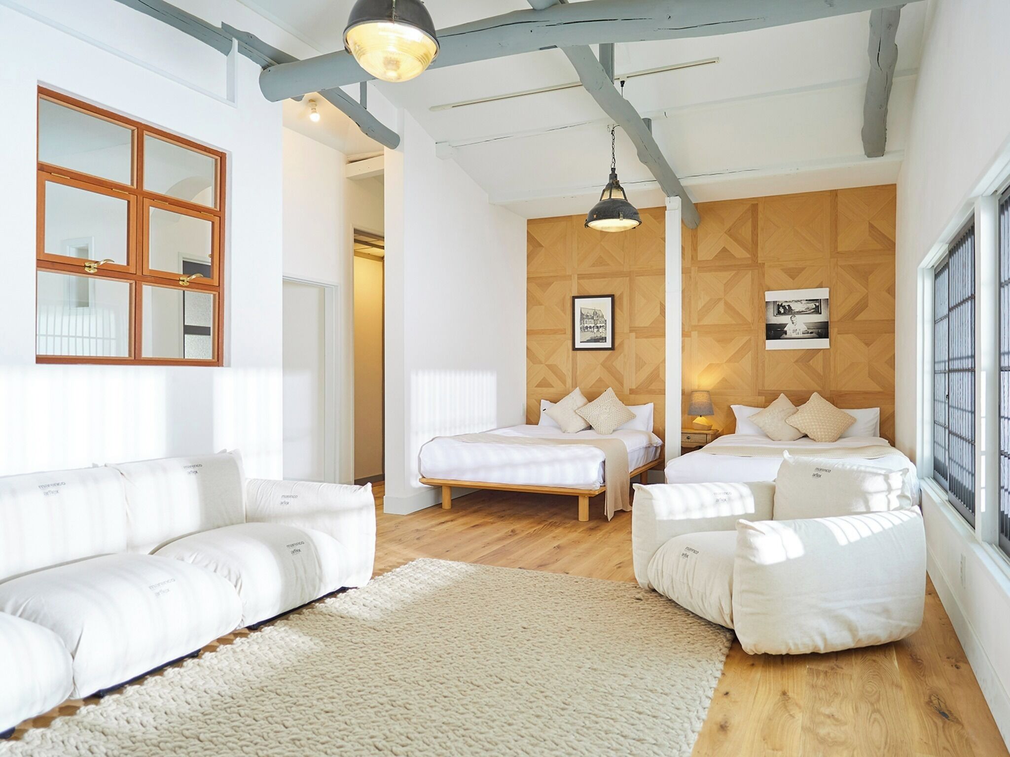 A modern bedroom with two beds, a white sofa, wooden flooring, and a large window.