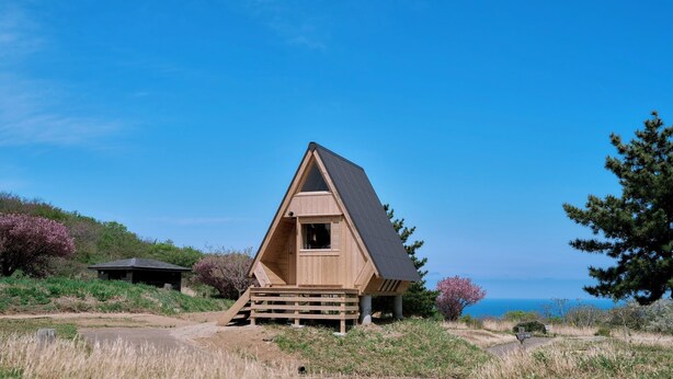 Una cabaña de madera con techo triangular, ubicada en una colina cubierta de hierba y con vista despejada al océano.