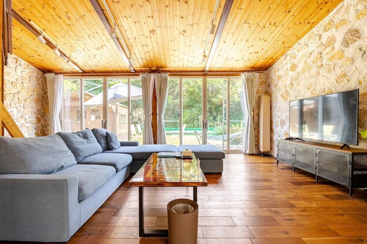 Una sala de estar moderna con techo de madera, paredes de piedra, un sofá gris, una mesa de centro de vidrio y un televisor sobre un soporte de metal.