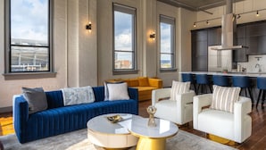 Una sala de estar moderna con un sofá azul, sillones blancos, una mesa de centro redonda y una zona de cocina con taburetes de bar.