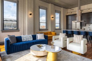 A modern living room with a blue sofa, white armchairs, a round coffee table, and a kitchen area with bar stools. - Luxury Penthouse | Rooftop Deck Best Street in OTR (Cincinnati)