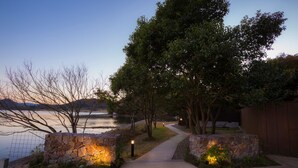 A pathway leading to a stone wall with lights, surrounded by trees and a body of water. - Mizube No Hanare Calme (Kyotango)