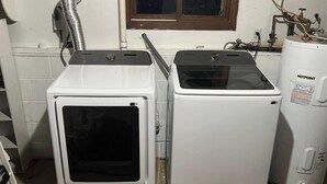 Deux machines à laver blanches se trouvent dans une pièce avec une fenêtre, un chauffe-eau et un conduit de ventilation.