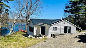 Une petite cabane avec un toit en métal, une allée en gravier et un lac en arrière-plan.