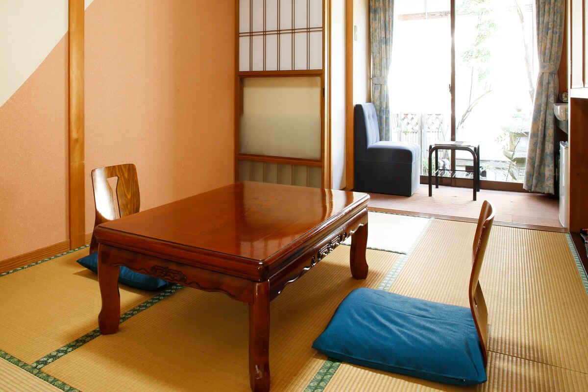 A traditional Japanese room with tatami flooring, a low wooden table, a blue cushion, and a sliding door leading to a balcony.