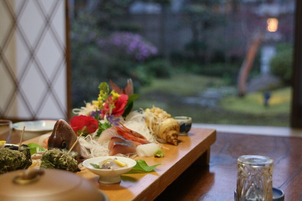 A wooden table with various seafood dishes and a small bowl of soup, set against a window with a view of greenery.