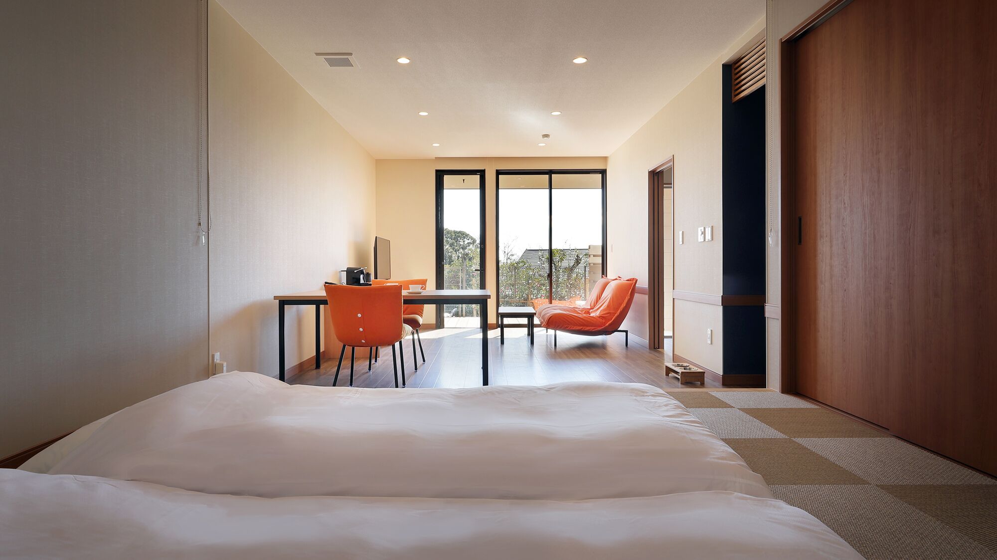 A hotel room with a bed, a desk, an orange chair, and a view of the outdoors.