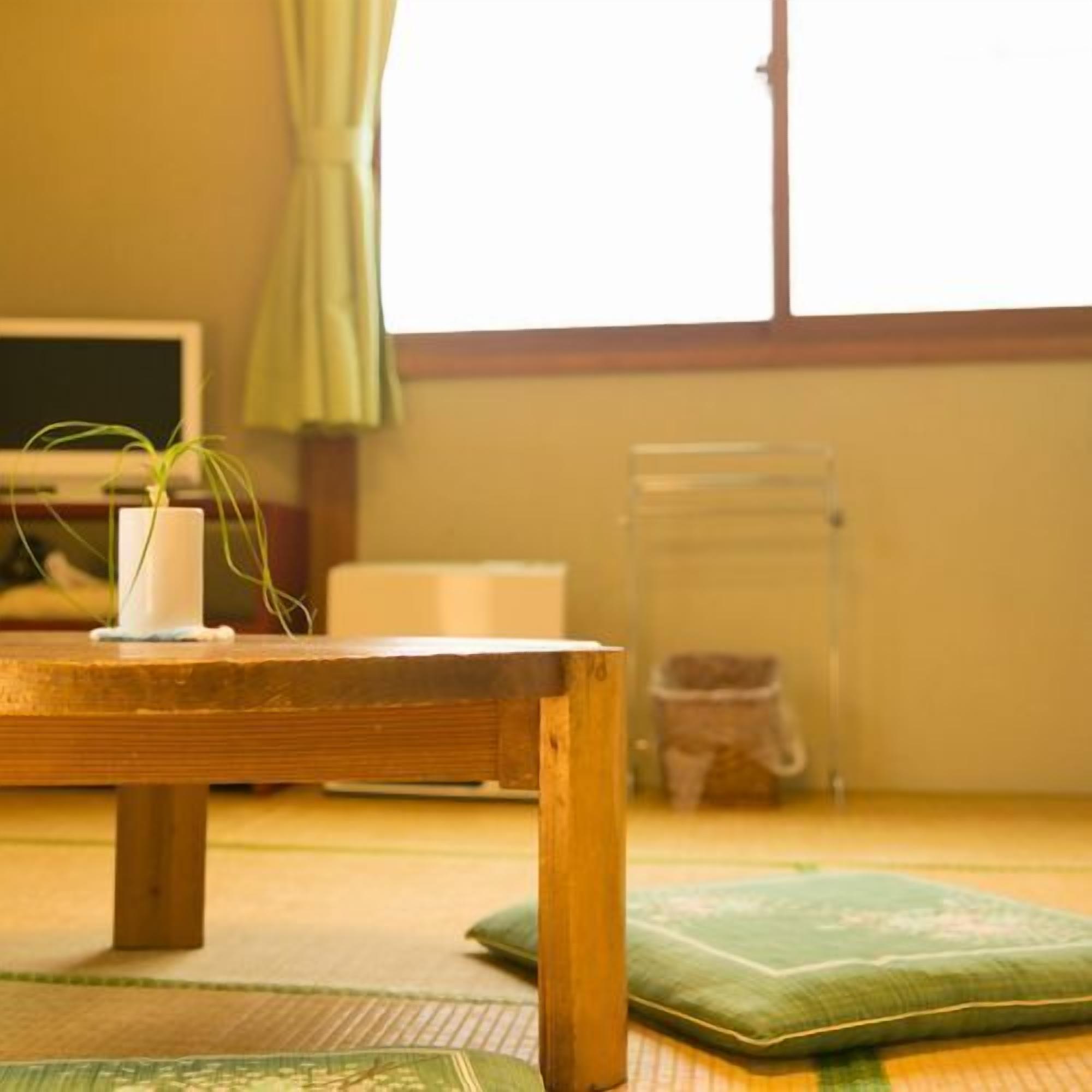 A room with tatami flooring, a wooden table, a green cushion, a white vase with a plant, and a window with curtains.