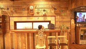 A child standing at a wooden bar counter with a television displaying a snowy scene, surrounded by wooden walls and various potted plants. - Tabi Yado Rantan (Shinano)