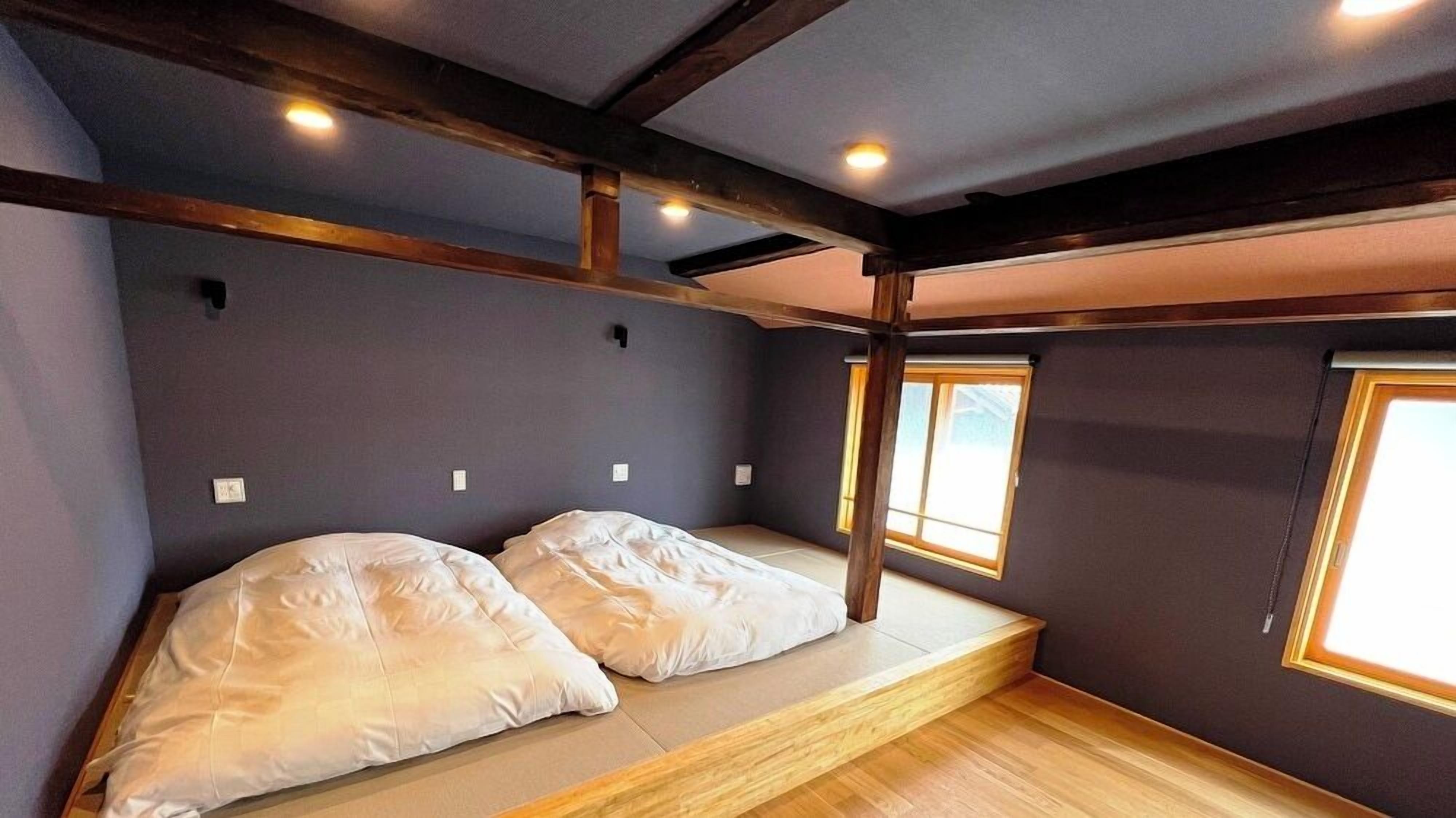 Habitación pequeña con literas, vigas de madera y dos ventanas.