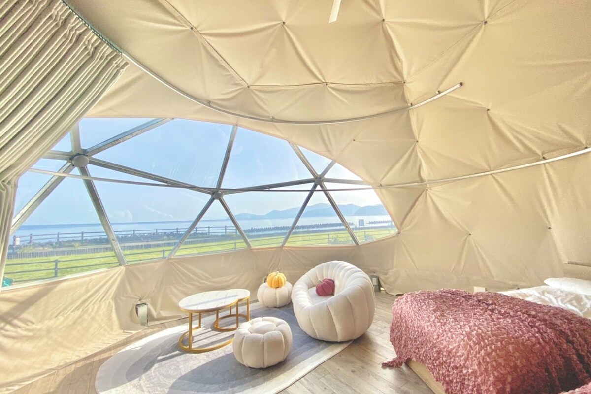 A cozy interior with a large window, a round table, two ottomans, a bed, and a view of the outdoors.