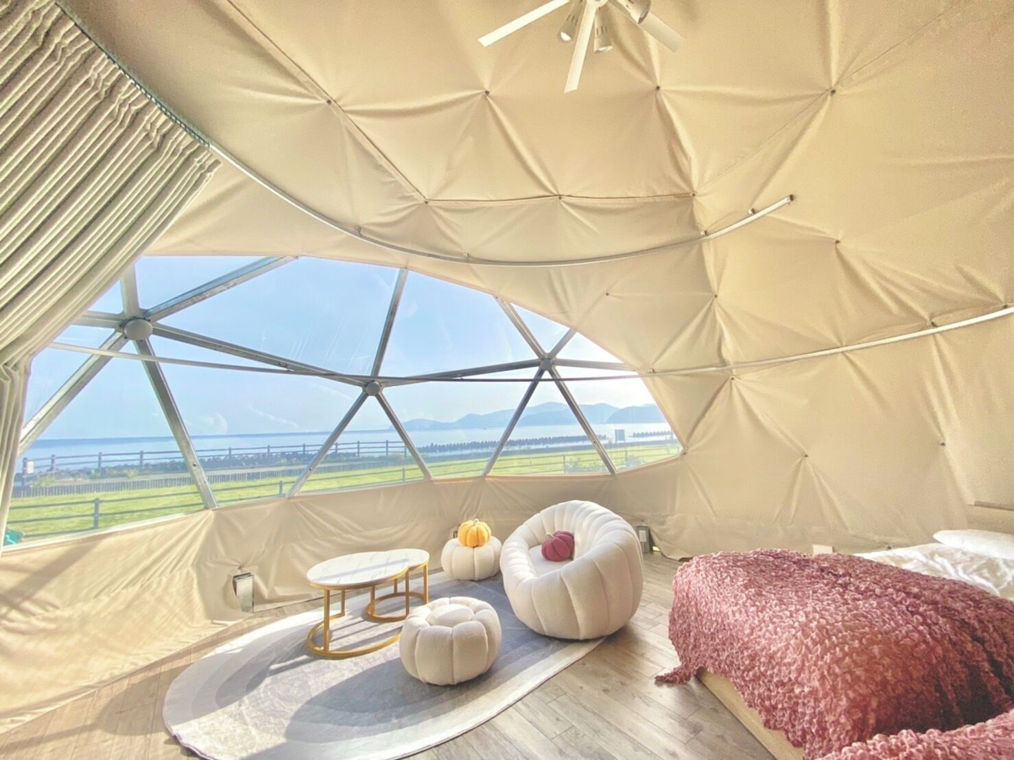 A cozy interior with a large window, a round table, two ottomans, a bed, and a view of the outdoors.