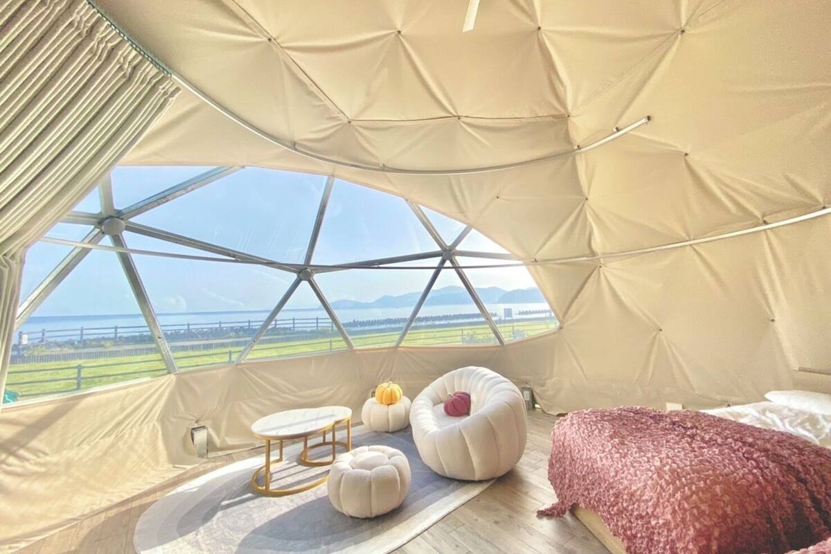 A cozy interior with a large window, a round table, two ottomans, a bed, and a view of the outdoors.