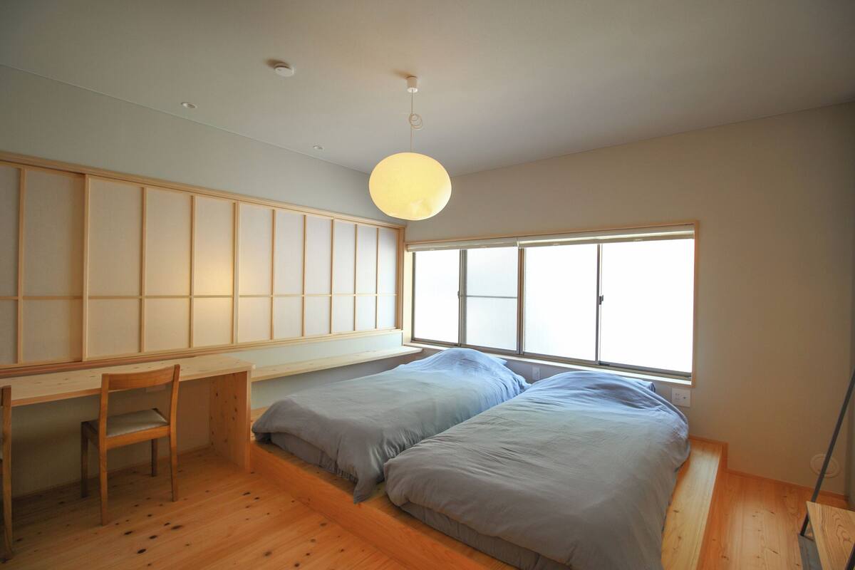 A room with a desk, two beds, a chair, and a large window.