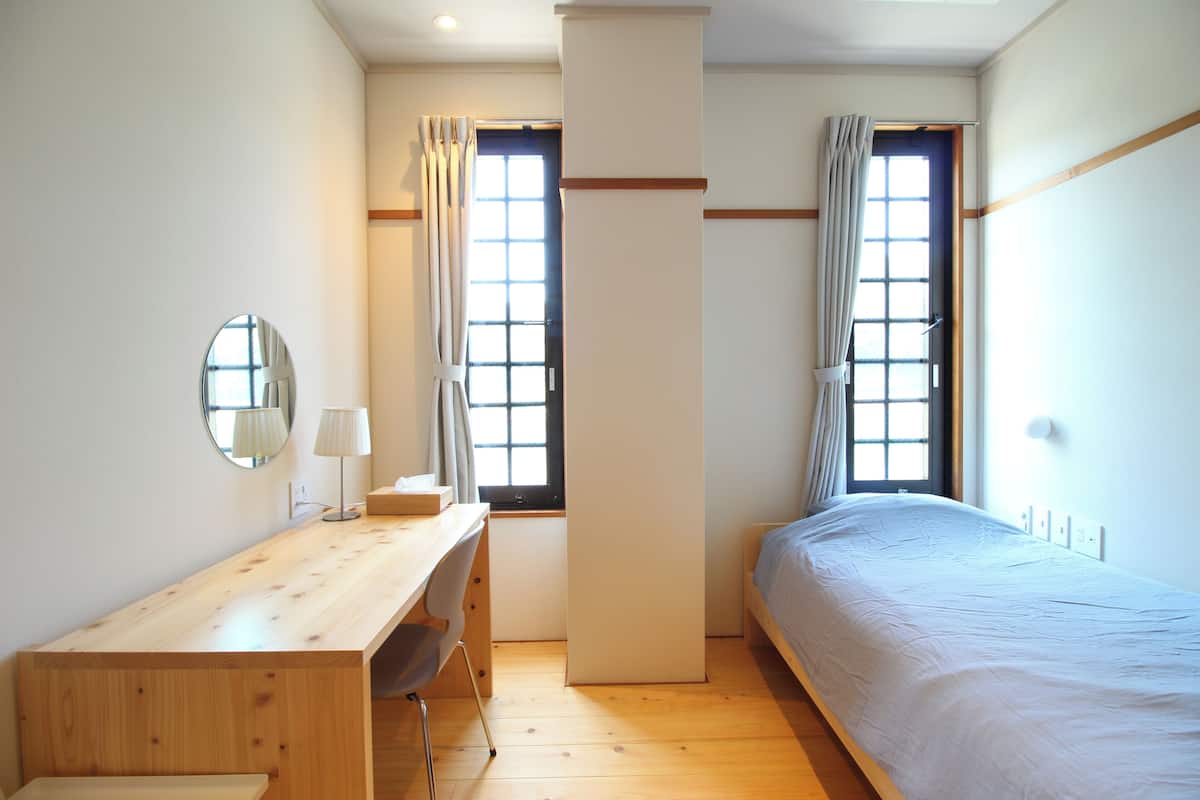 A bedroom with a wooden desk, a bed, a chair, and two windows with curtains.