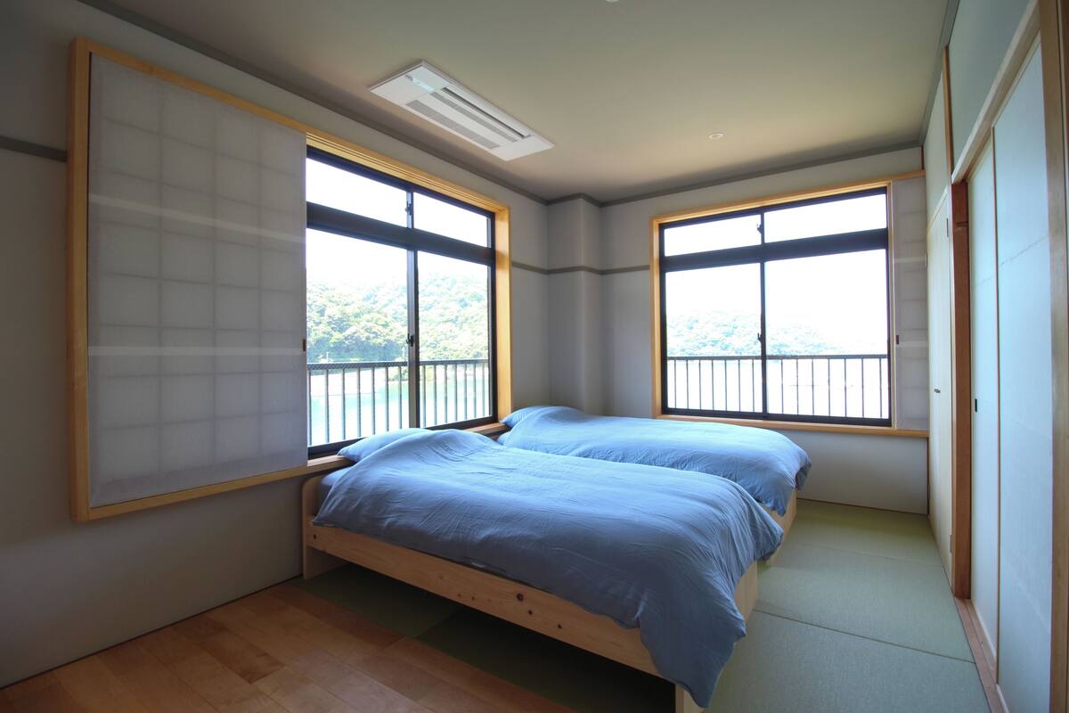 A room with two beds, large windows, a ceiling fan, and a view of the outdoors.