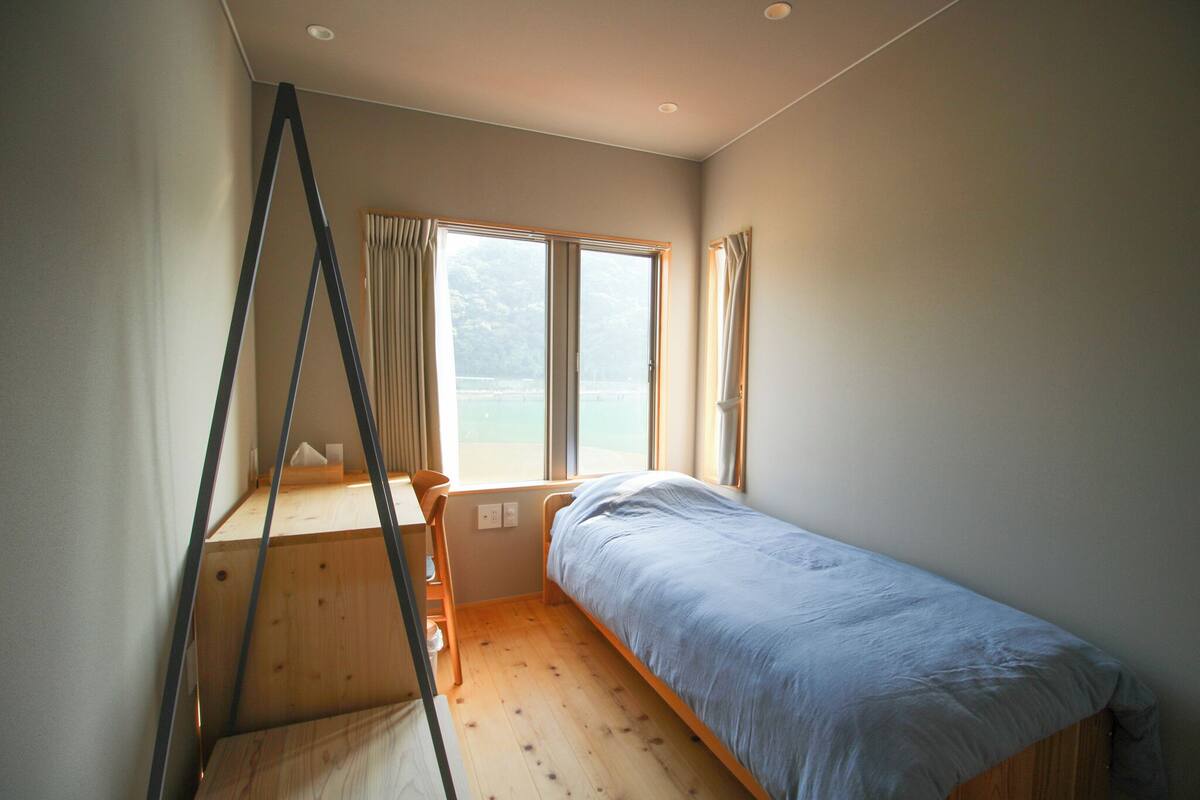 A small, single-bed room with a desk and chair, large windows, and a view of the outdoors.