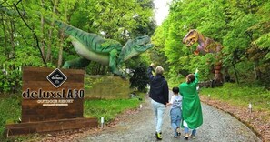 Un letrero de madera con el texto 'deluxsLABO' y 'OUTDOOR RESORT', rodeado de follaje y estatuas de dinosaurios.