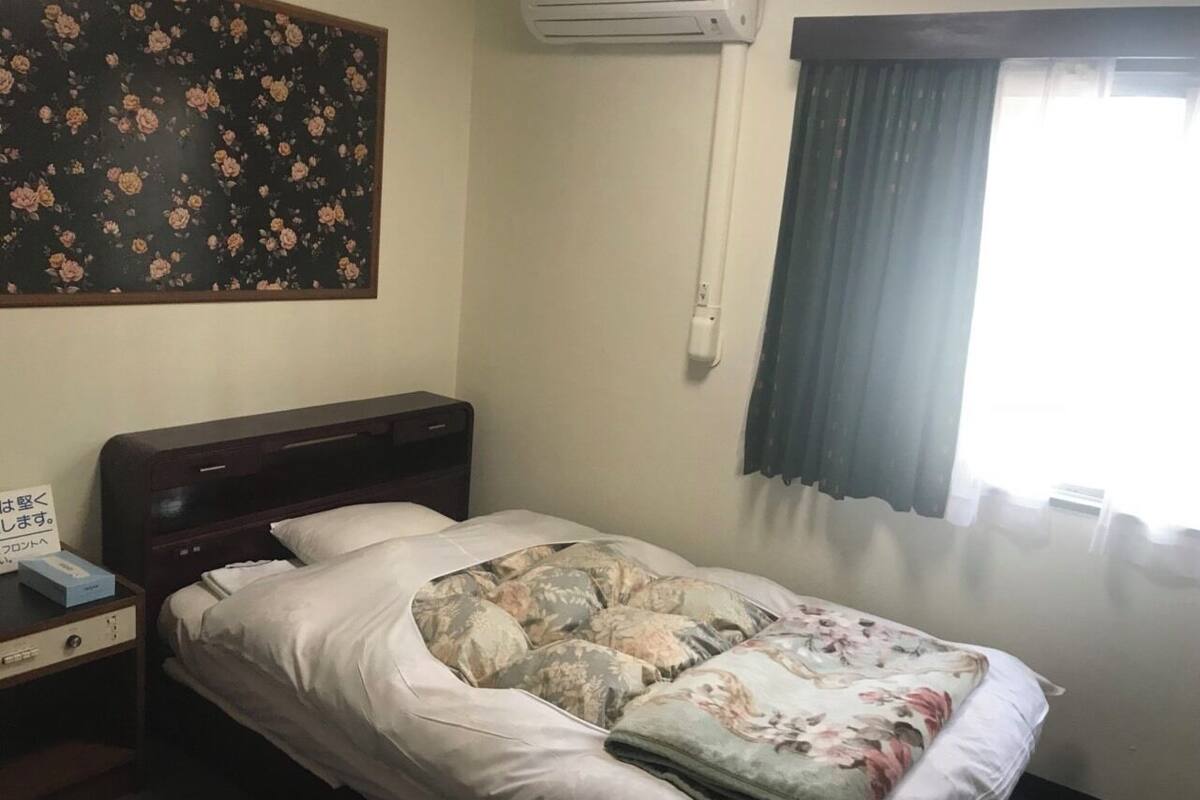 Een eenpersoonsbed met een bloemenprint sprei, een nachtkastje met een telefoon en een doos, een wandairconditioner en een raam met gordijnen.