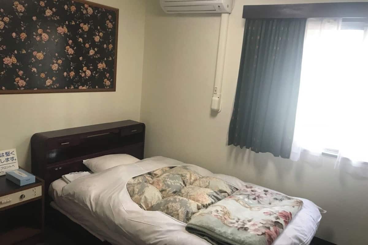 A single bed with a floral bedspread, a nightstand with a phone and a box, a wall-mounted air conditioner, and a window with curtains.