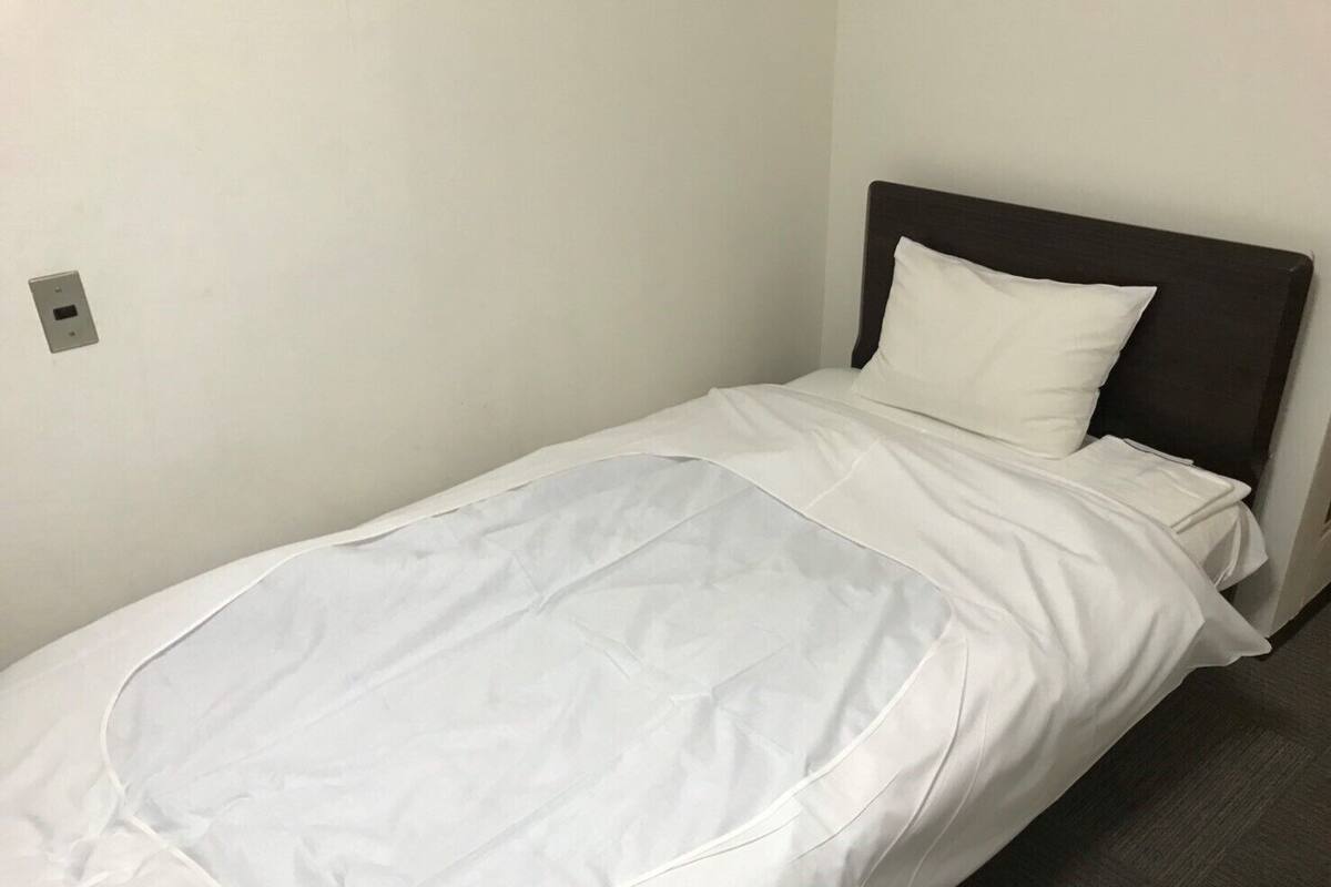 Een eenpersoonsbed met een witte matras en kussen, een donker hoofdbord en een effen wand met lichtschakelaar.