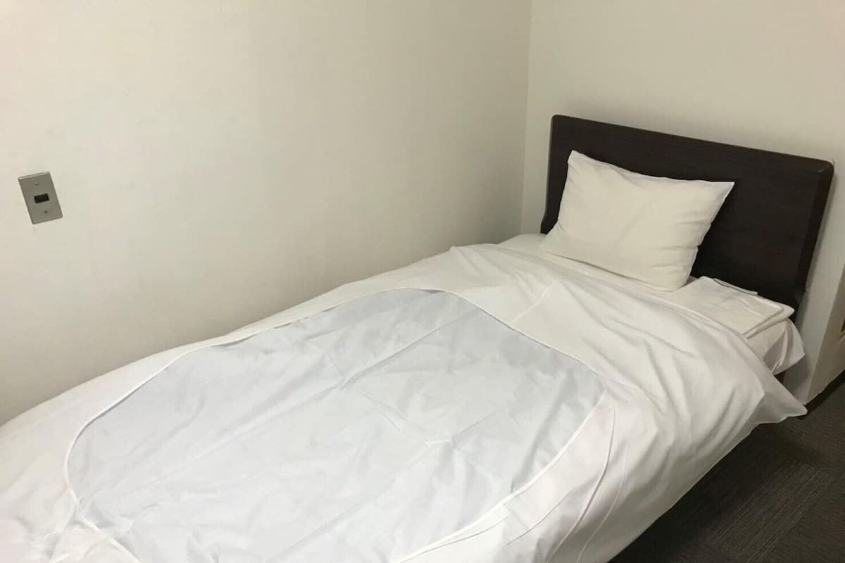 A single bed with a white mattress and pillow, a dark headboard, and a plain wall with a light switch.