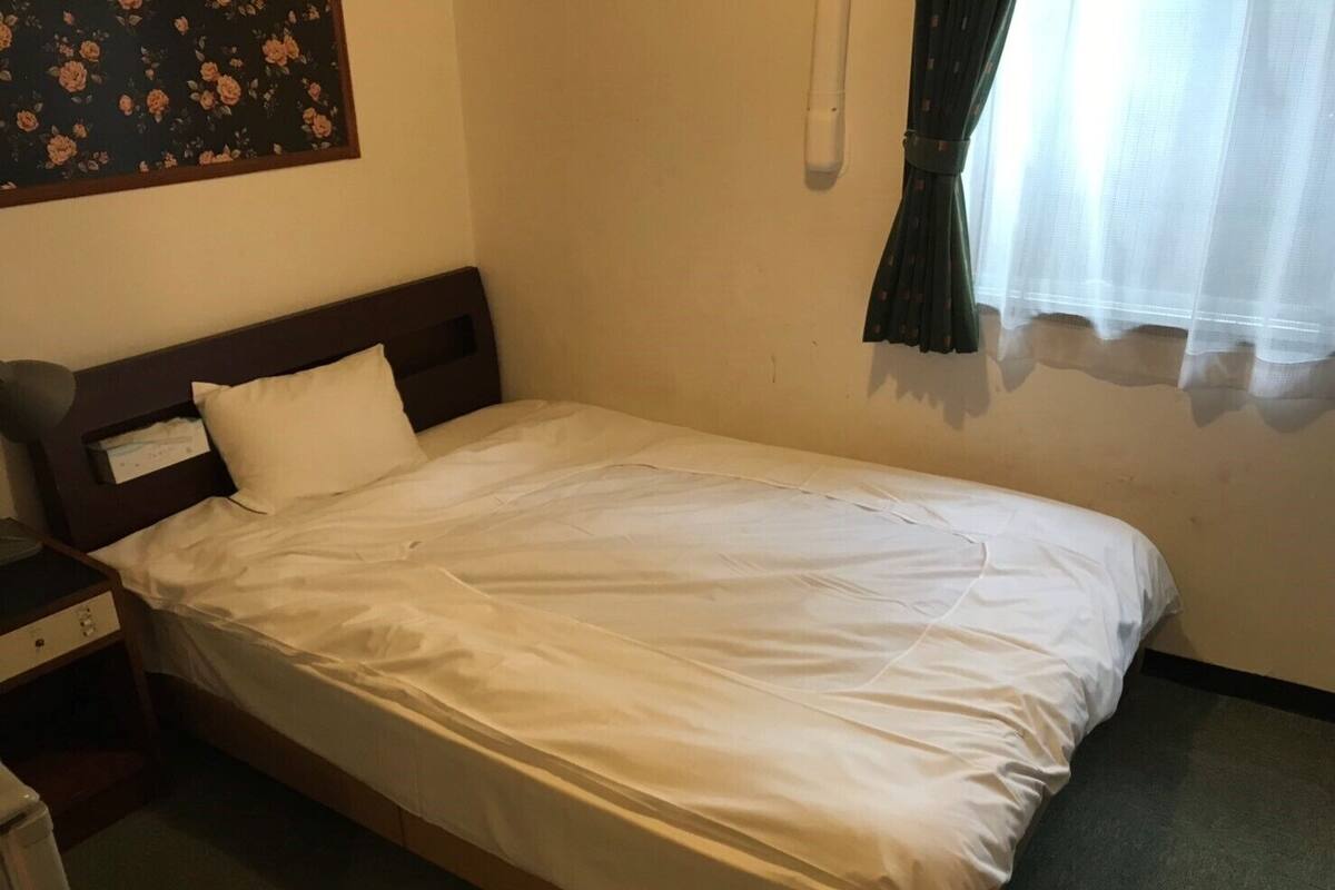 Een enkel bed met witte beddengoed, een nachtkastje, een wandlamp en een bloemenprint aan de muur.