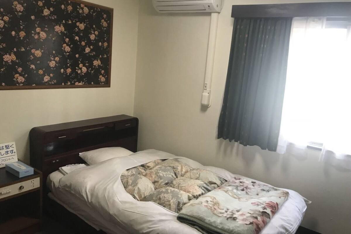 Een eenpersoonsbed met een bloemenprint dekbedovertrek, een nachtkastje met een telefoon en een doos, een wandairconditioner en een raam met gordijnen.