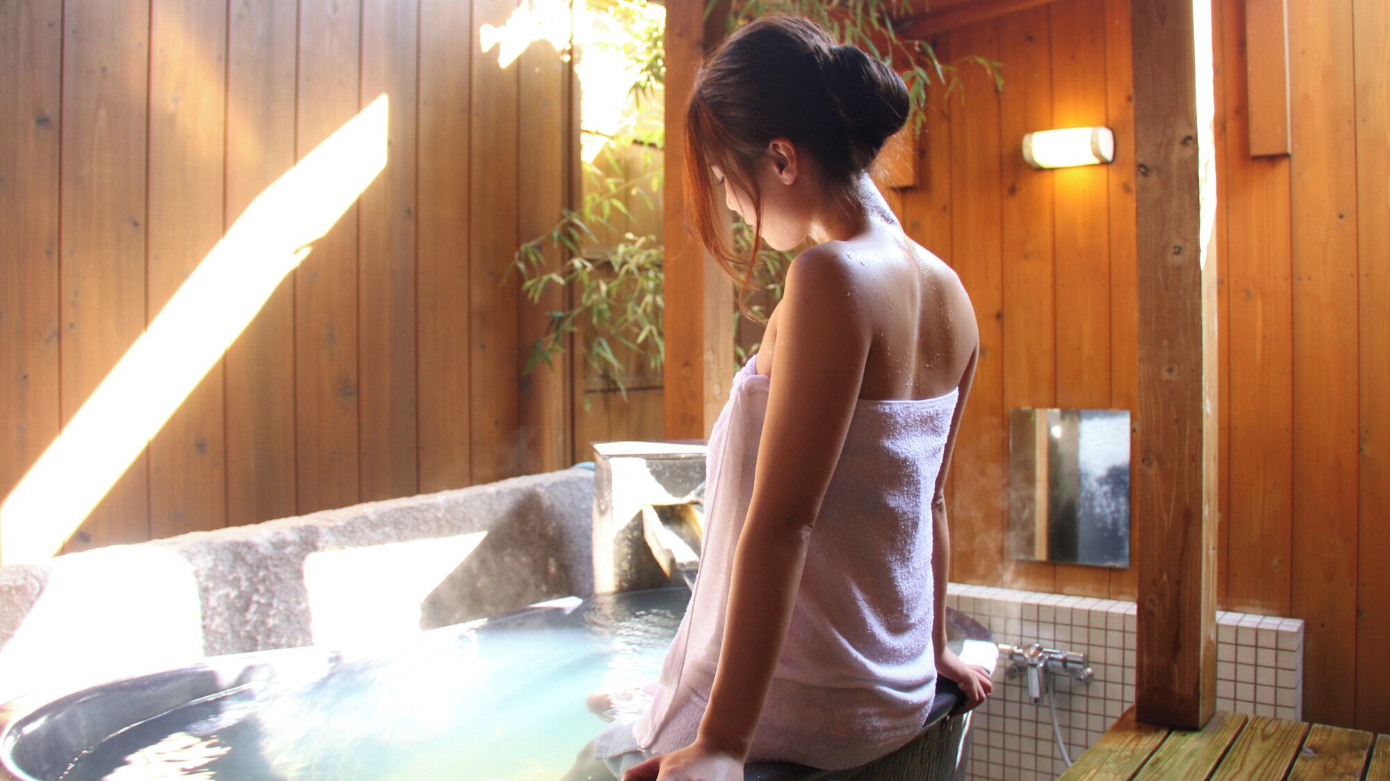 A person in a towel standing by a hot tub in a wooden room with a tiled wall and a small table.