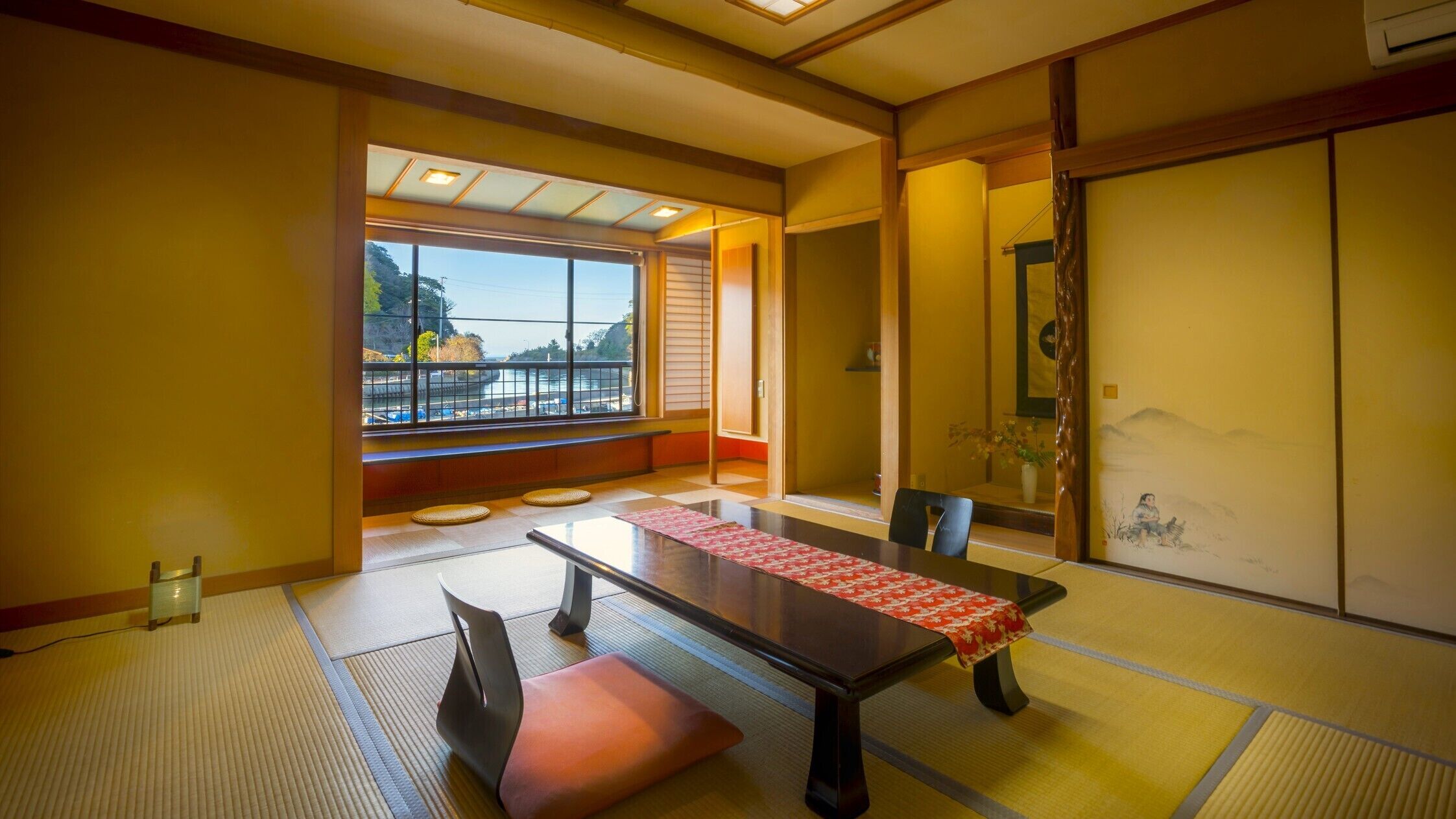 Habitació japonesa tradicional amb terra de tatami, una taula baixa amb un corredor vermell i una porta corredissa amb una pintura. Es veu un pàrquing a través de grans finestres.