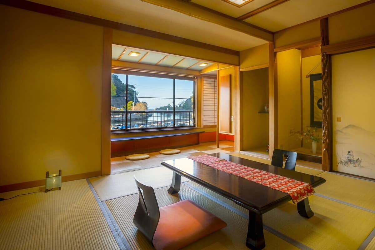 A traditional Japanese room with tatami flooring, a low table with a red table runner, a sliding door with a painting, and a view of a parking lot through large windows.