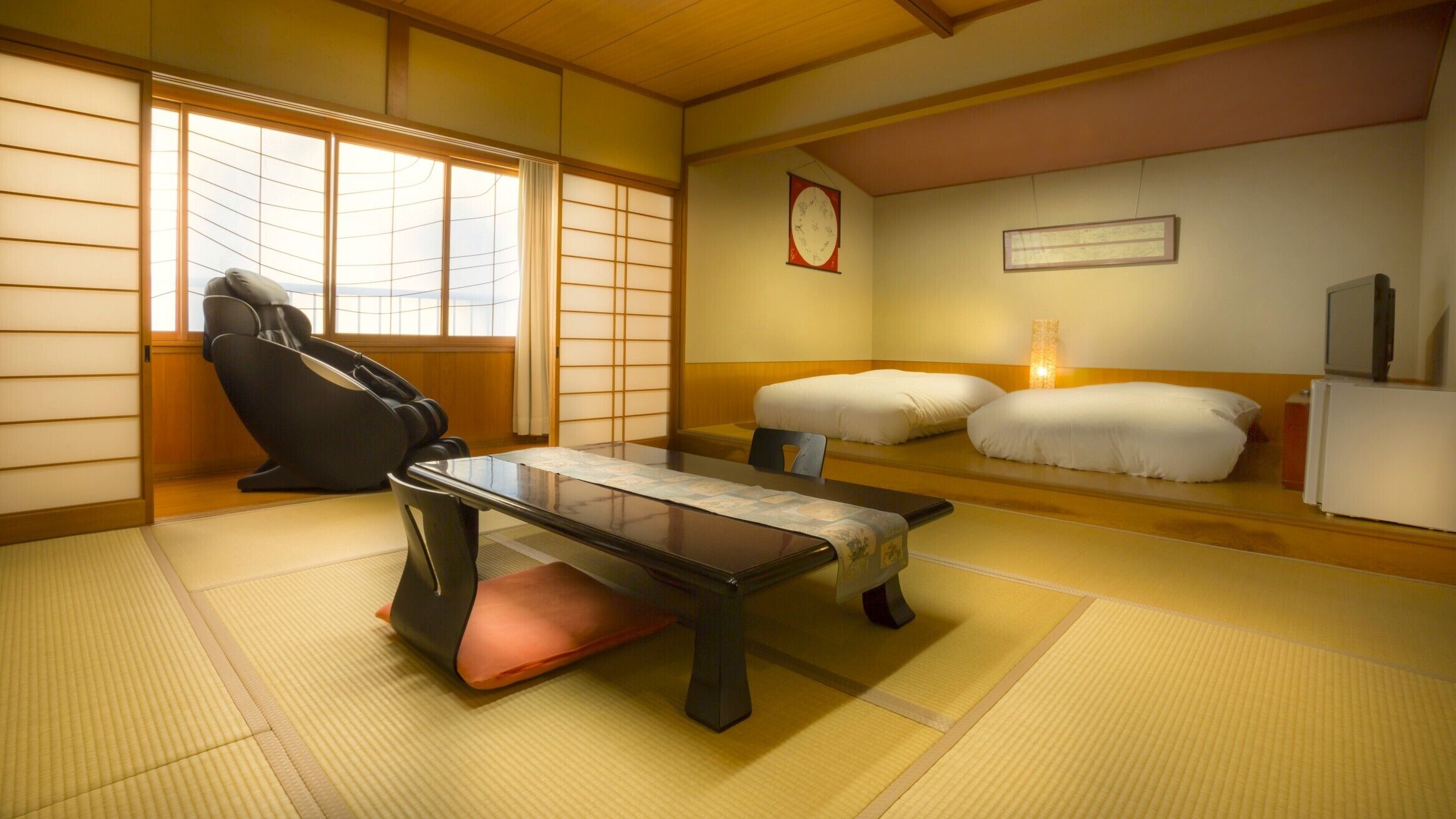 A traditional Japanese room with tatami flooring, a massage chair, a low table, and a bed with pillows.