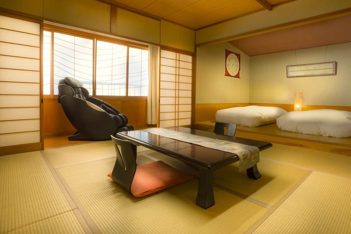 A traditional Japanese room with tatami flooring, a massage chair, a low table, and a bed with pillows.