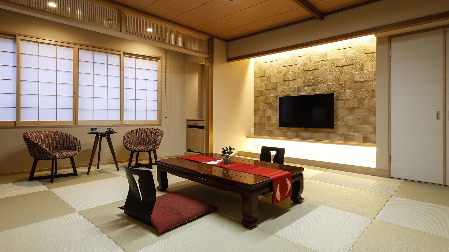Habitació tradicional japonesa amb sòl de tatami, una taula baixa, dues cadires, un televisor fixat a una paret de pedra i portes corredisses.