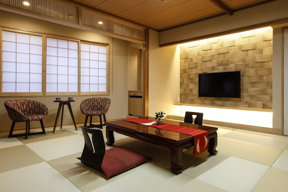 A traditional Japanese-style room with tatami flooring, a low table, two chairs, a TV mounted on a stone wall, and sliding doors.