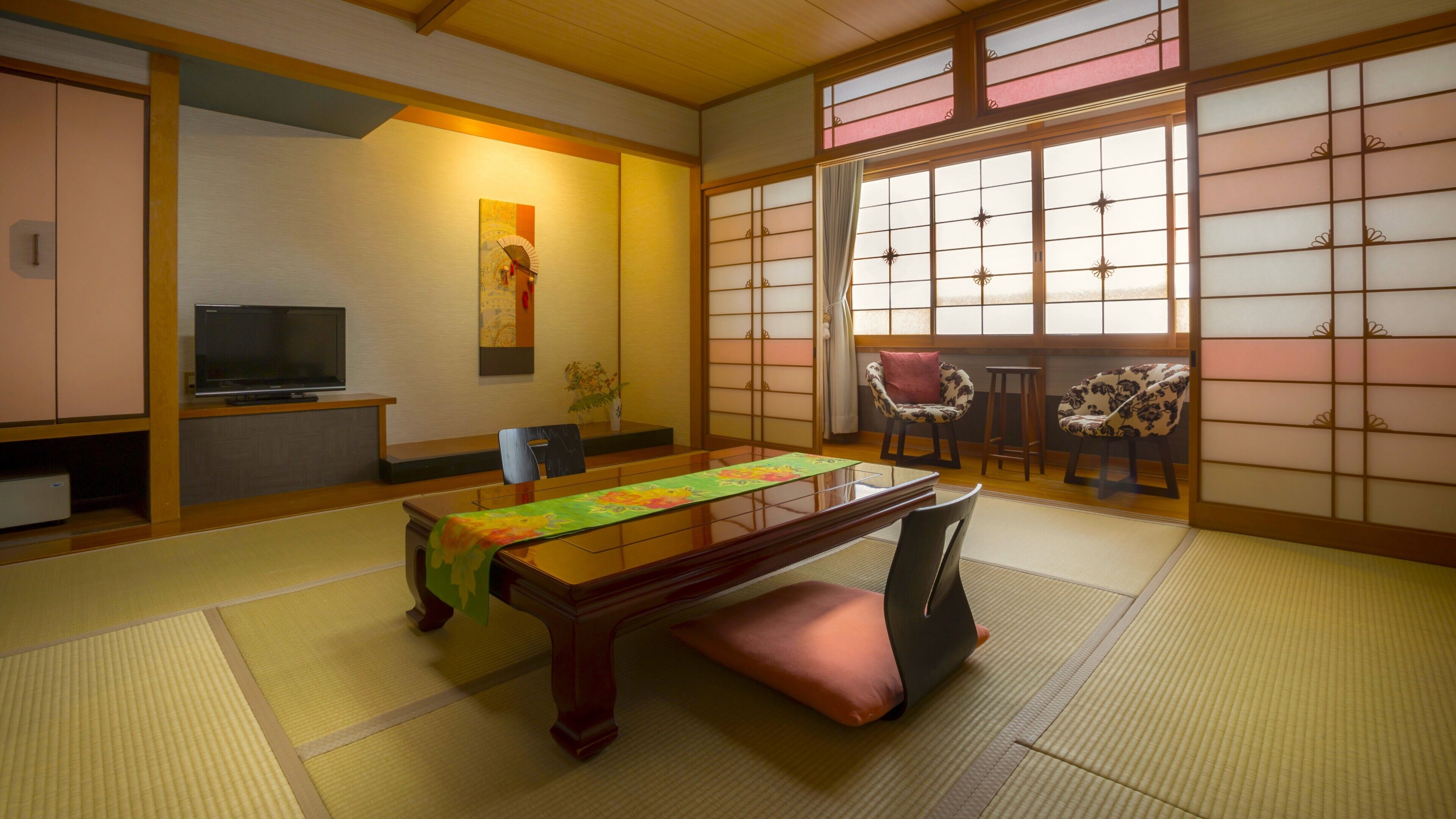 A traditional Japanese room with tatami flooring, a low table with a green tablecloth, a television on a stand, and sliding doors with a view of the outside.