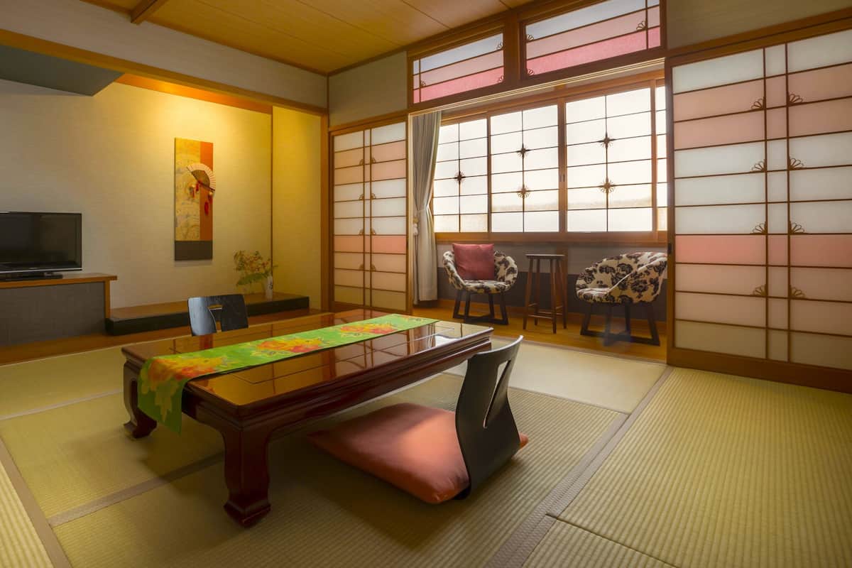 A traditional Japanese room with tatami flooring, a low table with a green tablecloth, a television on a stand, and sliding doors with a view of the outside.