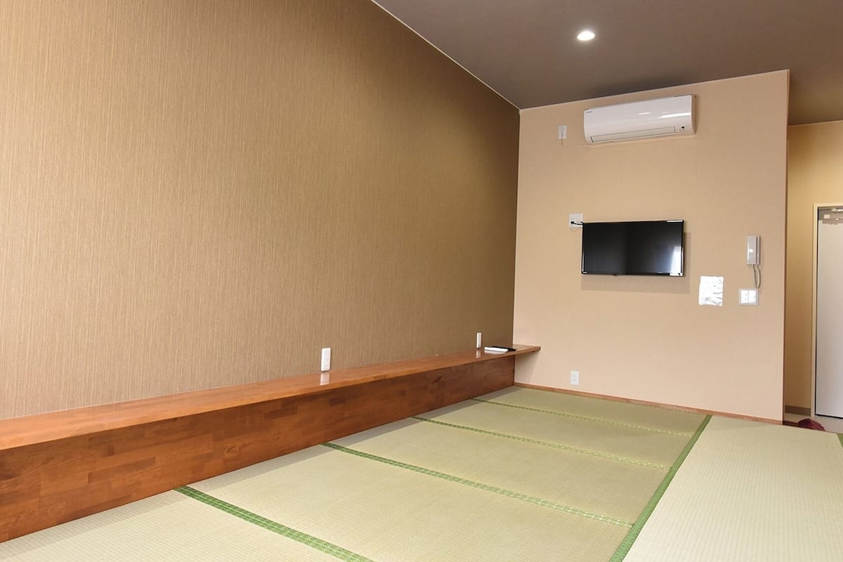 Ein Zimmer mit Tatami-Matten, einer hölzernen Bank, einem Wand-Fernseher und einer Wand-Klimaanlage.